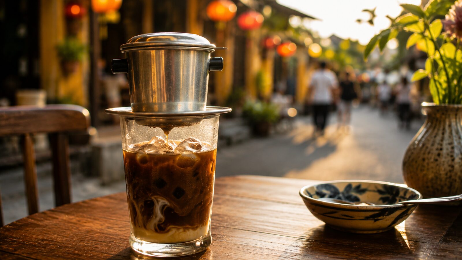 A glass of Vietnamese iced milk coffee with a metal phin filter on top, a lantern-lit street behind.