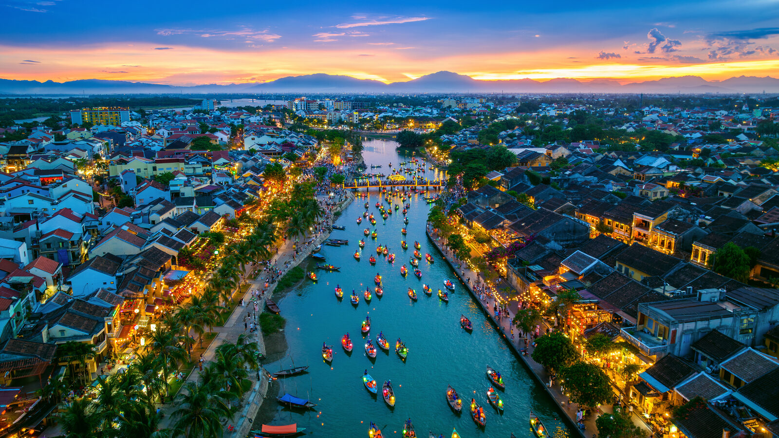 Hội An at golden hour, lantern-lit boats on the Thu Bồn river and ochre shophouses under an orange sky.