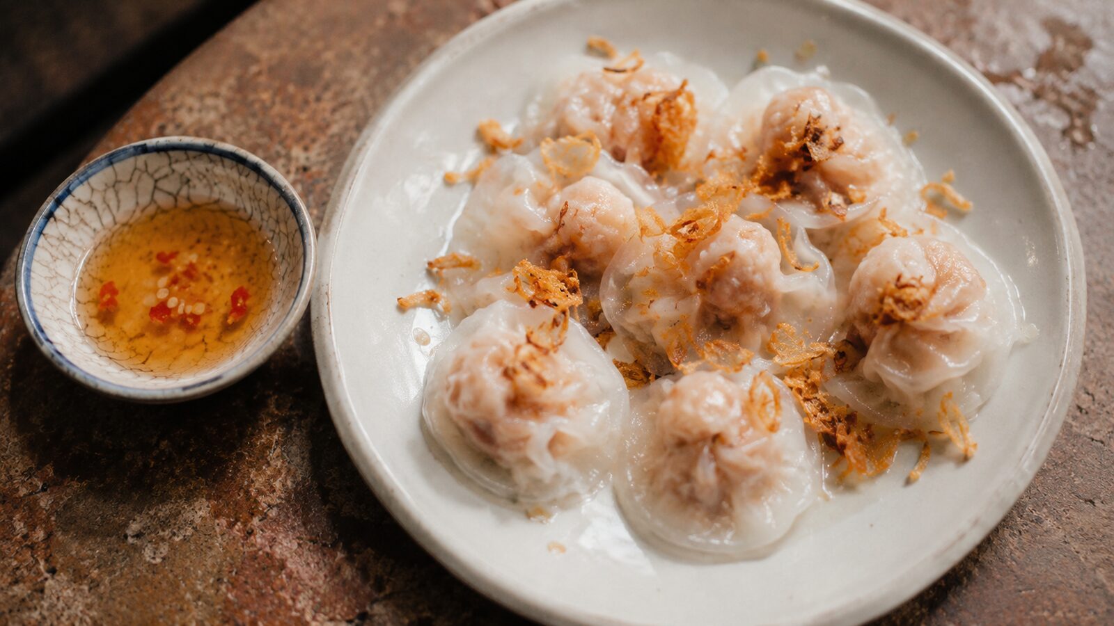 A plate of translucent Hội An white rose dumplings topped with crispy fried shallots, with dipping sauce.